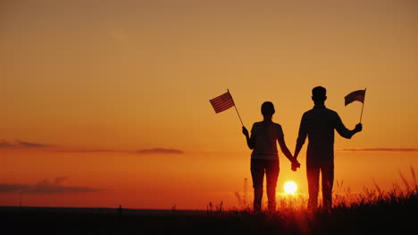 Una-Joven-Pareja-Con-Las-Banderas-De-Los-Estados-Unidos-En-Sus-Manos-Admira-La-Hermosa-Atardecer-Indep