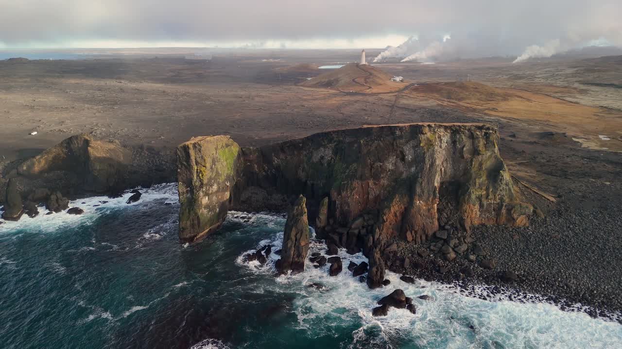 Aerial Approaching shot of Coastline in Reykjanes with crashing waves against rocky coast. Geothermal hot springs and rising steam in background. Iceland, Europe. Wide shot.