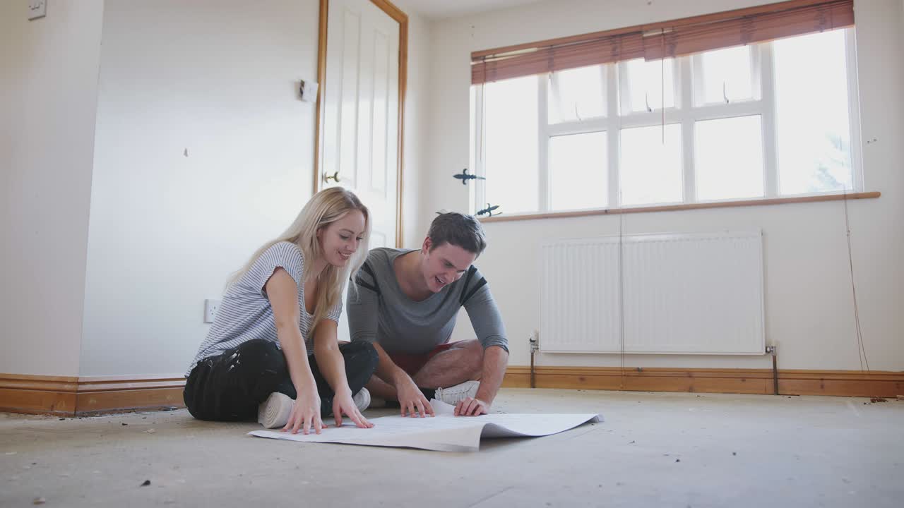 Pareja sentada en el suelo mirando planos de planta en una habitación vacía de su nuevo hogar.