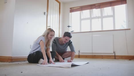 couple sitting on floor looking at floor plans in empty room of new home