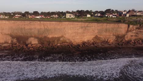 Aerial-trucking-flight-along-massive-cliffs,-waves-and-town-on-top-during-sunset-time