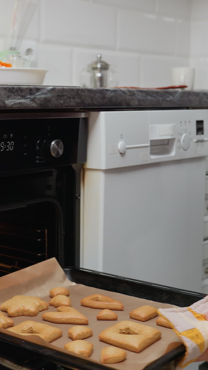 Signora che usa uno strofinaccio per togliere i biscotti cotti dal forno con una pentola sul piano di lavoro, biscotti su carta da forno, cucina ben illuminata, biscotti caldi e dorati appena sfornati