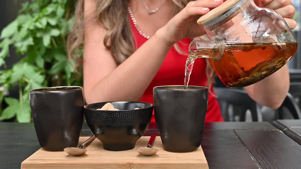 Brunette woman in a red dress pouring tea in a black cup from a glass tea infuser