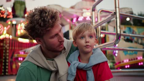 Shooting-in-the-reach,-a-father-with-curly-hair-in-a-Green-T-shirt-communicates-with-his-little-son,-a-blond-man-with-blue-eyes-in-a-red-T-shirt-against-the-backdrop-of-bright-attractions-with-bright-lanterns-and-light-bulbs-in-an-amusement-park.-Dad-and-son-hugging