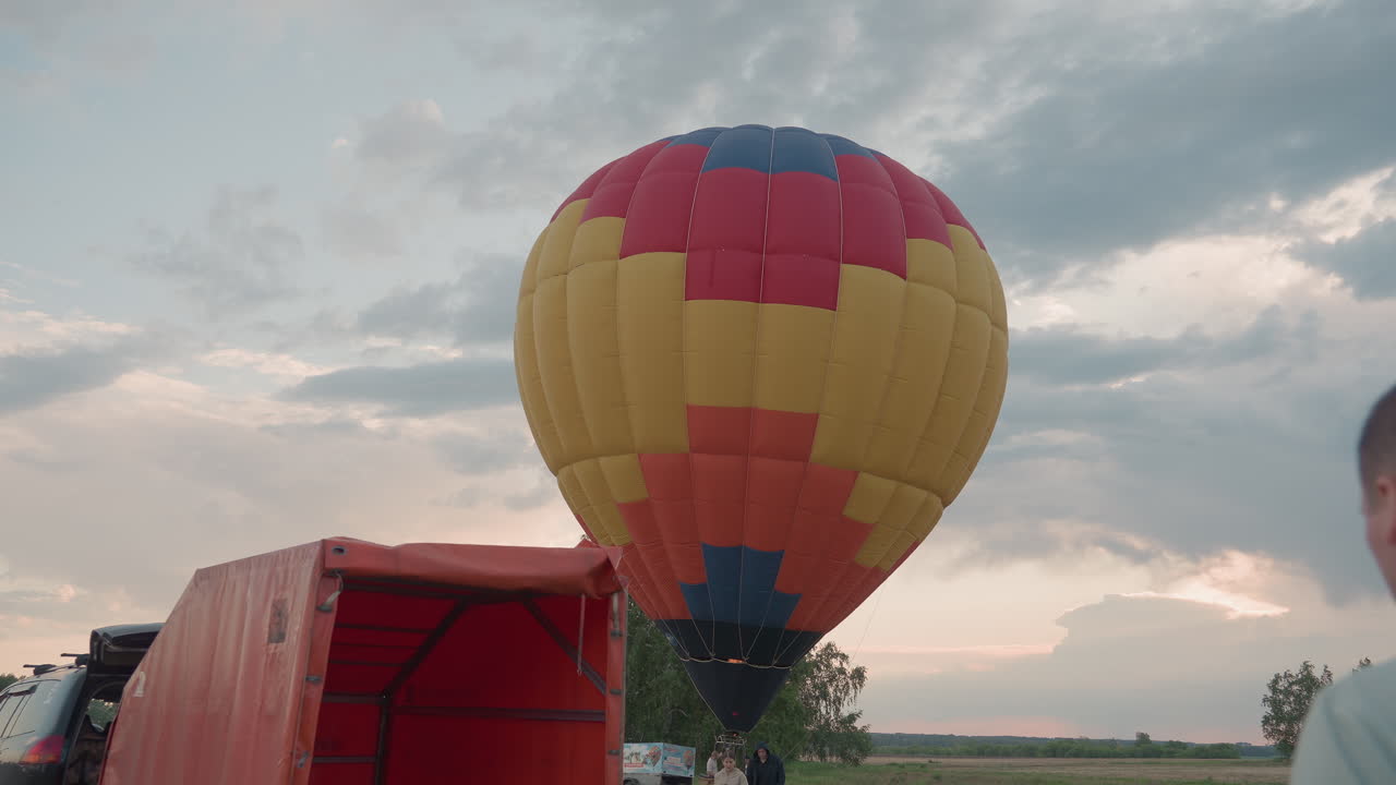 multicolor hot air balloon partially inflated standing adjacent to open trailer and suv in green field at sunset while crew carry basket toward launch site preparing ascent under dramatic cloud sky