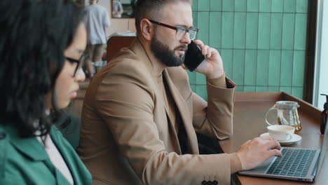 Hombre-De-Negocios-Hablando-Por-Teléfono-Y-Usando-Una-Computadora-Portátil-En-El-Café