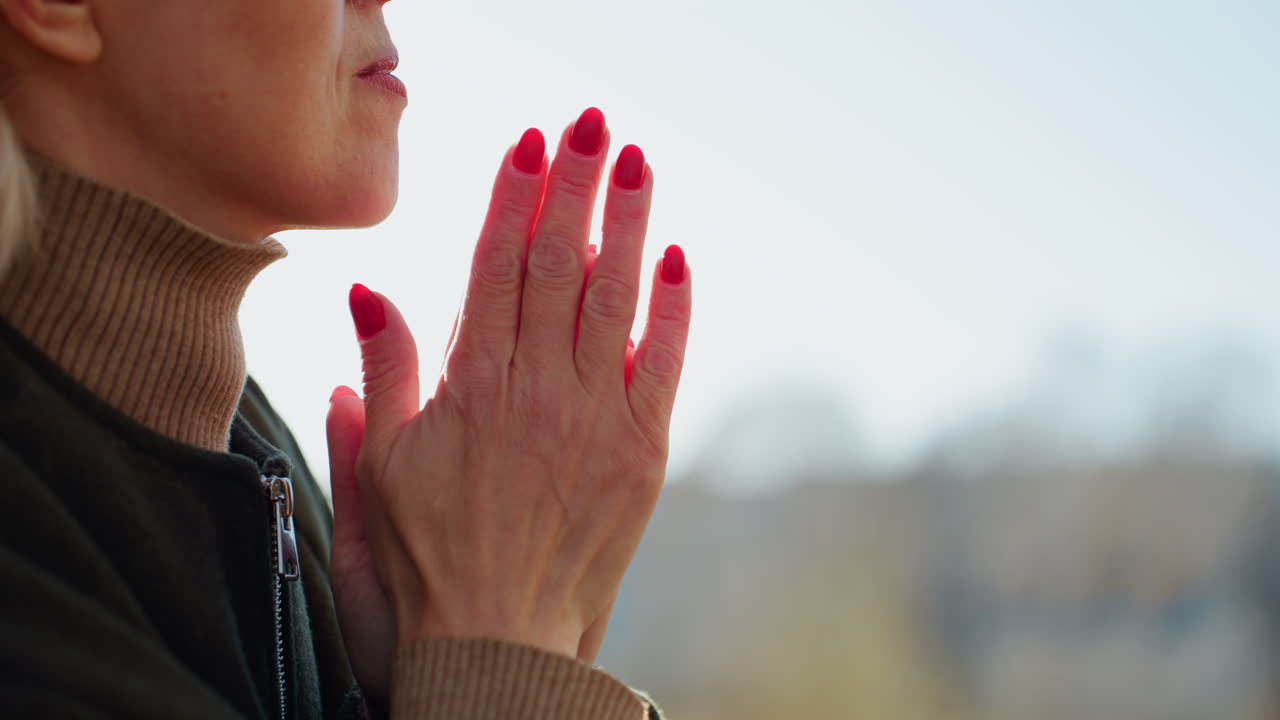 Side view of mature woman with blonde hair and red nails clasping hands together outdoors in sunlight, wearing warm clothing, expressing gratitude, reflection and emotional calmness