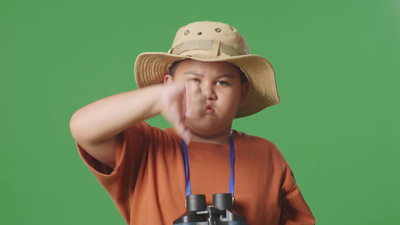 모자를 입은 아시아 관광 소년은 안경을 통해 쳐다본 후 엄지손가락을 아래로 표시합니다. 소년 연구자는 녹색 화면 배경에서 무언가를 조사합니다.