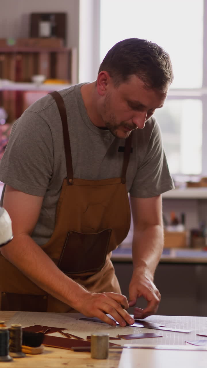 Bearded leathersmith cuts leather sheet with cutter at workplace in home studio. Handcrafting master makes product in traditional way. Tailor at work