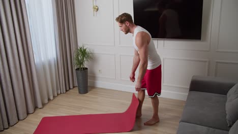 happy male athlete with a short haircut and beard at home in a sports summer suit lays out a mat and warms up before playing sports inside the house. side view of a happy blond man warming up in a white t-shirt and red shorts before starting his exercise routine at home