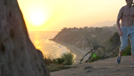 young fit shirtless man watching a golden sunset after a workout and run on the edge of a cliff above the pacific ocean in santa barbara, california