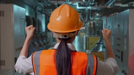 close up back view of a female engineer with safety helmet raising her hands celebrating while working in engine control room, work of electrical generators
