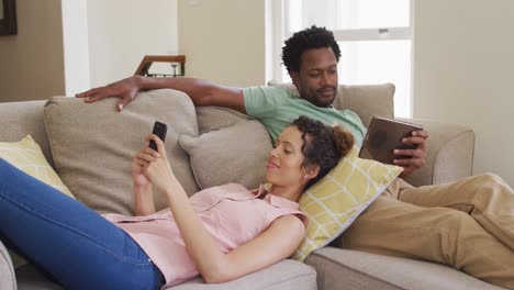 happy biracial couple relaxing on sofa with smartphone and tablet