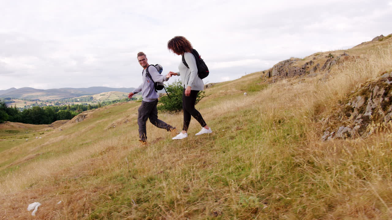una pareja de adultos jóvenes sonrientes caminando por una colina tomados de la mano durante una caminata por la montaña