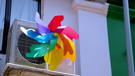 a vibrant multicolored pinwheel spins rapidly beside a building, mounted on an air conditioning unit, under bright natural sunlight with a clear blue sky