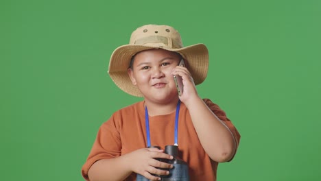niño turista asiático con un sombrero y binoculares hablando en teléfono inteligente mientras está de pie en el fondo de la pantalla verde. niño investigador examina algo, concepto de aventura de turismo de viaje, de cerca