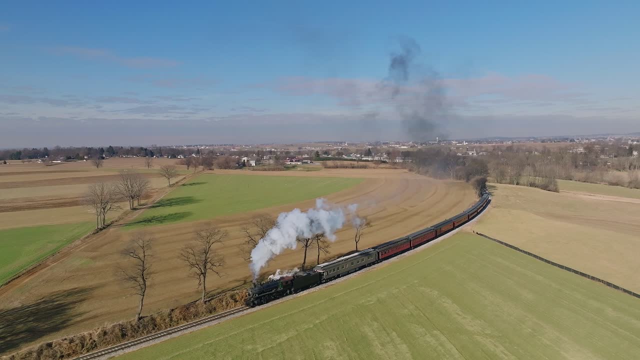 una vista aérea en cámara lenta, de un tren de pasajeros de vapor que se acerca, alrededor de una curva, soplando humo, mientras viaja por el campo, en un día soleado de invierno, en un día soleado de invierno