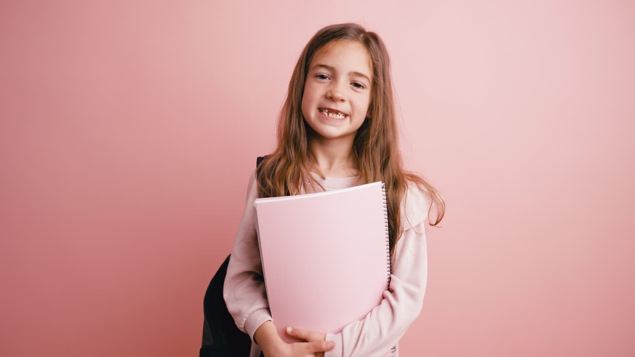 Glad ung skolflicka som håller en anteckningsbok och ler på rosa bakgrund
