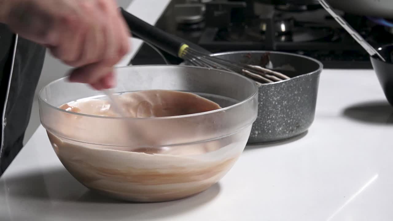 Watch as a skilled cook blends cream smoothly in a glass bowl, preparing for a delightful dessert in a cheerful kitchen filled with natural light