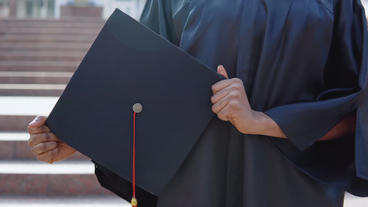 Square master's hat vertically in the hands of an African-American female university graduate on the background of the stairs from the outside.