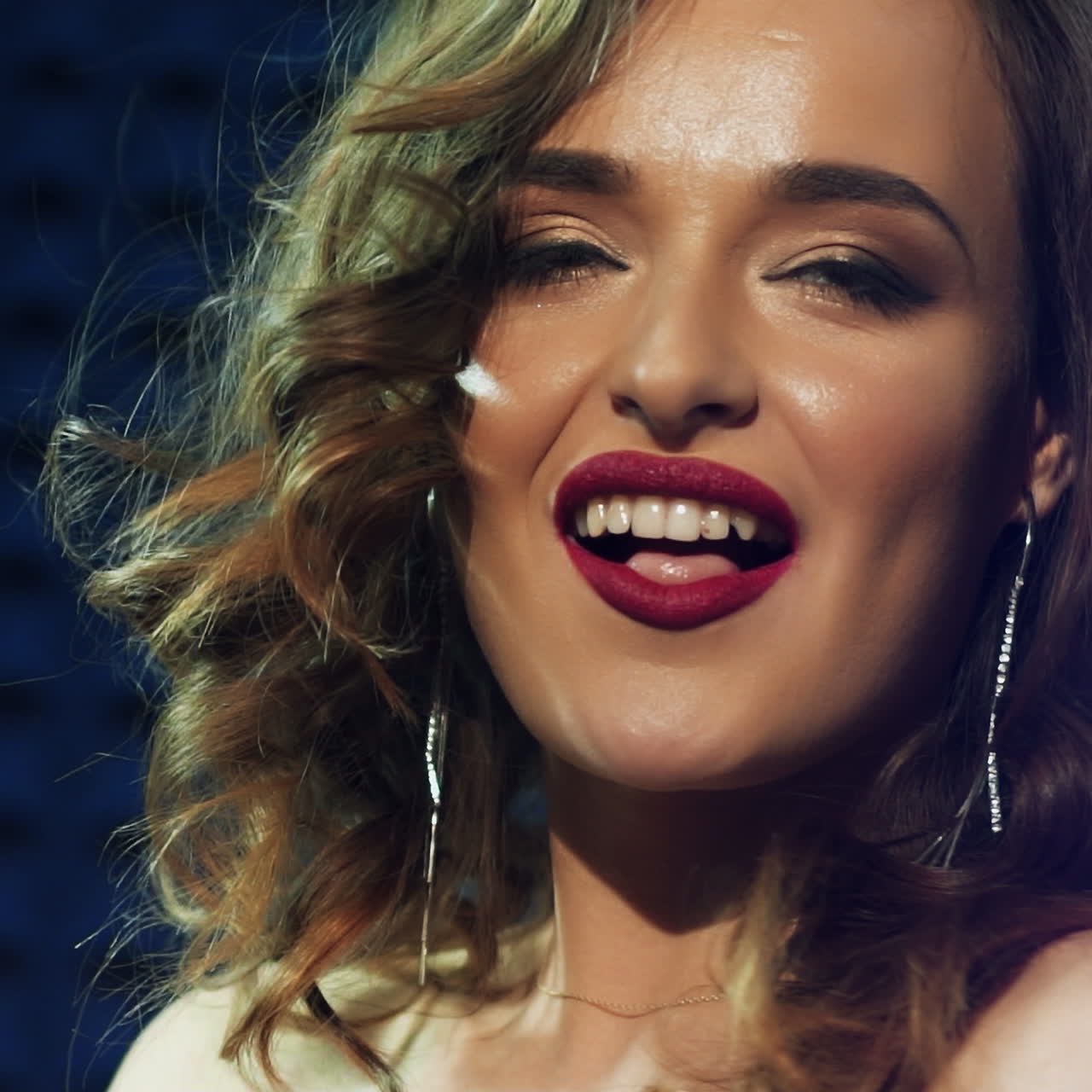 Slow motion of gorgeous woman with bright make-up looking at camera and smiling isolated on blue background. Portrait of a lovely singer with long wavy hair. Close-up