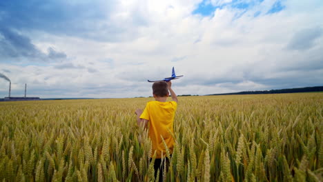 little boy plays on factory background. child in yellow t-shirt walking on wheat field and holds toy plane in his hand in summer.