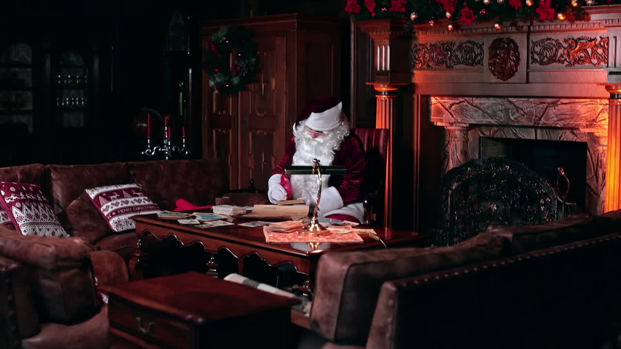Hardworking Santa Claus writing letters and Christmas cards. Adorable decorated interior with old furniture and fireplace backdrop.