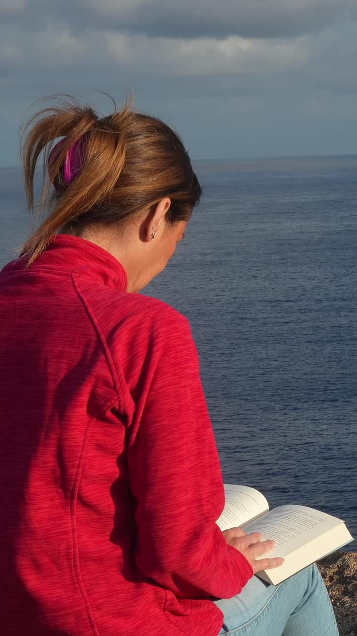 Donna che legge un libro seduta su una scogliera con vista sull'oceano in una giornata nuvolosa e calma