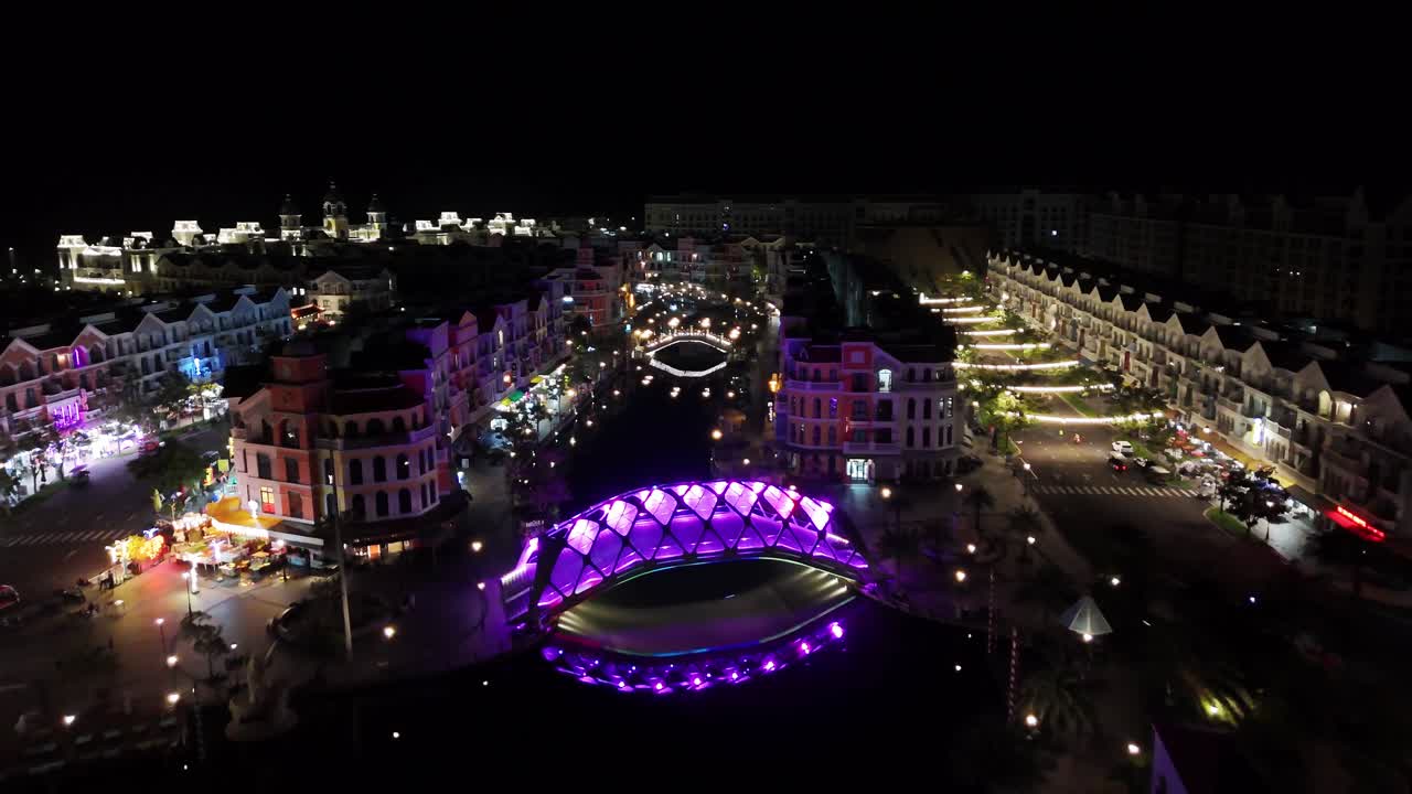 Nighttime aerial video showcases a brightly illuminated urban waterfront in Phu Quoc featuring European-style buildings along a canal crossed by illuminated pedestrian bridges and lively evening walks