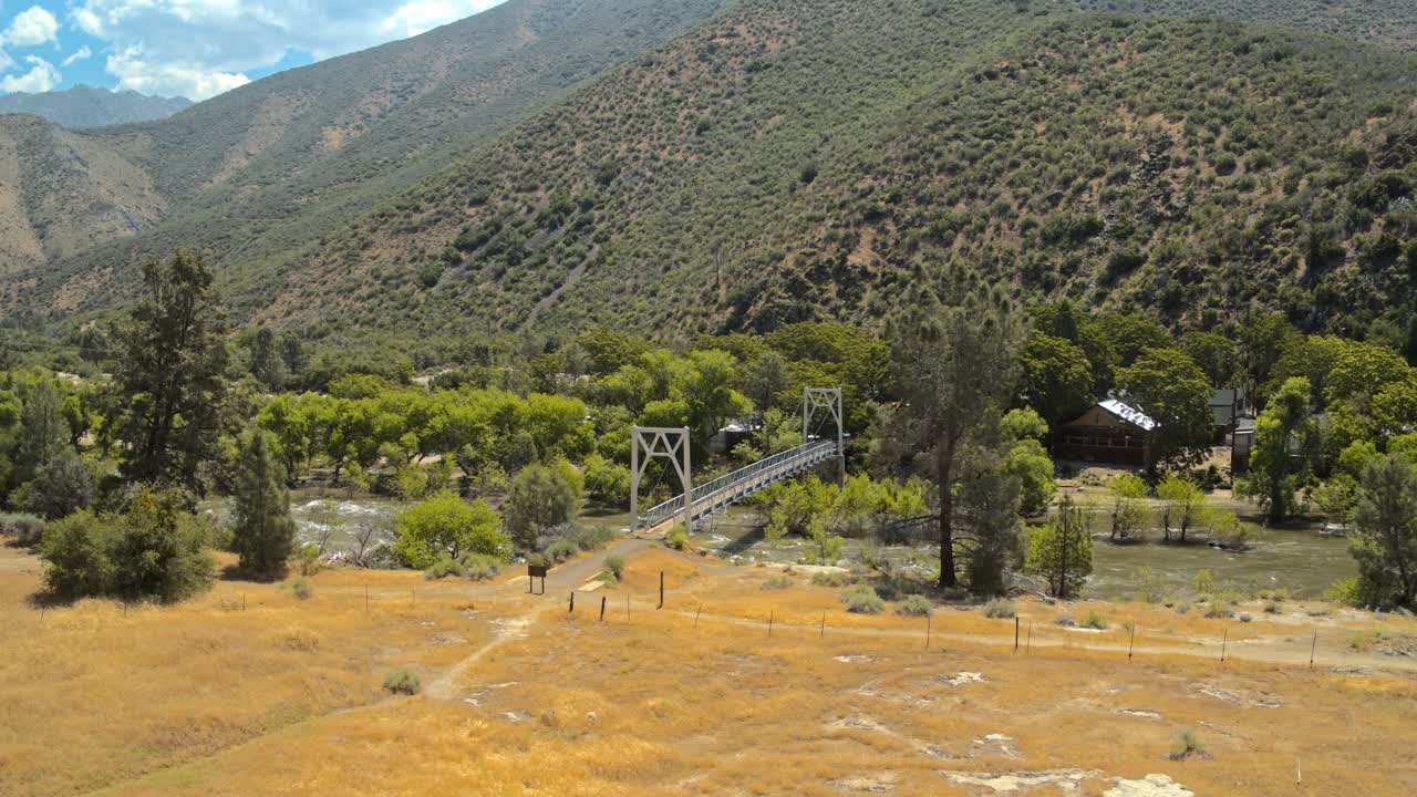 Aerial shot of a river flowing under a suspension bridge by a cabin.