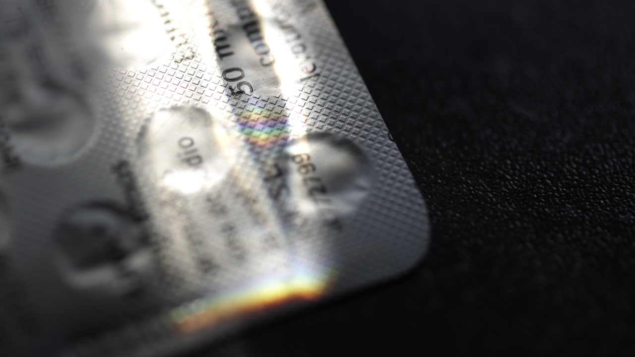 Rainbow colored light illuminating pharmaceutical blister pack, casting vibrant reflections across dark textured surface, highlighting intricate packaging details and medication design