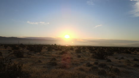 golden sunlight descends over a vast desert landscape, illuminating the low shrubs and shadows as it sinks beneath the horizon, creating a peaceful atmosphere.