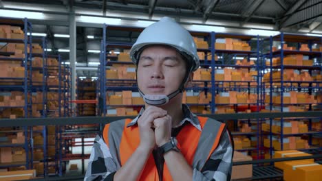 primer plano de ingeniero masculino asiático con casco de seguridad reza por algo mientras está de pie en el almacén con estantes llenos de mercancías de entrega