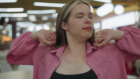 lady in pink dress looking bored takes a deep breath and stretches her neck by tilting her head side to side while placing her hands on her neck, in the background, a man in black passes by