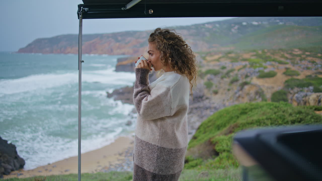 Tender traveller drinking hot beverage looking on wonderful gloomy seascape.