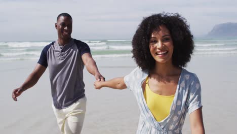 smiling african american couple walking and holding hands at the beach
