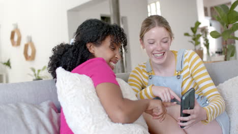 amigas adolescentes felices y diversas hablando y usando teléfonos inteligentes en el sofá en casa, en cámara lenta