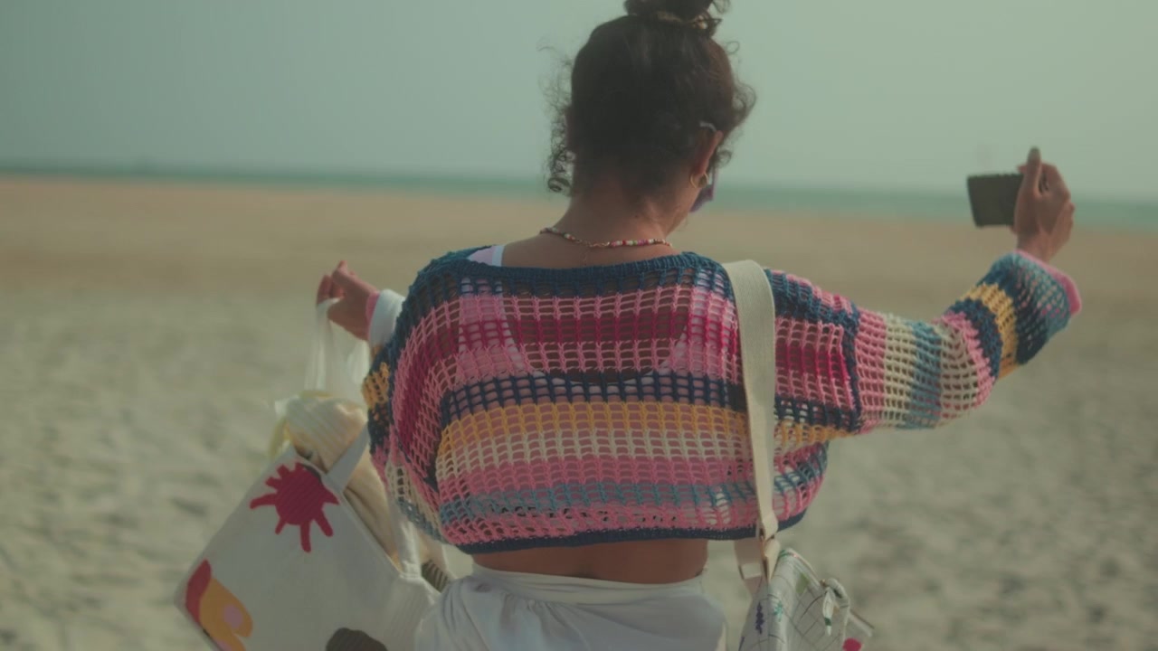 Donna con borse si scatta un selfie su una spiaggia sabbiosa, indossando un maglione colorato e occhiali da sole
