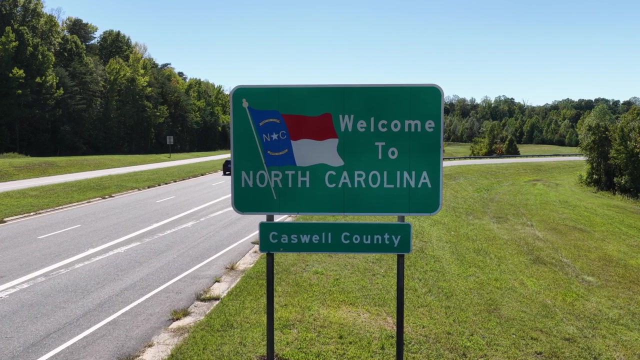 Un'auto passa sulla corsia vicina accanto al cartello di benvenuto in North Carolina per la contea di Caswell, con l'autostrada che curva attraverso una fitta foresta su entrambi i lati. Veduta aerea