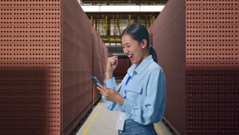 side view of asian female professional worker with her smartphone with red brick packed in stacks are stored, she raises her fist up with screaming goal after check on the smartphone