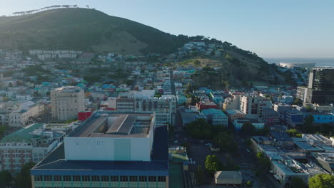 Fly-over-tall-buildings-and-heading-towards-residential-Bo-Kaap-borough.-Rows-of-coloured-low-houses-and-trees-on-top-of-Signal-Hill-in-background.-Cape-Town,-South-Africa