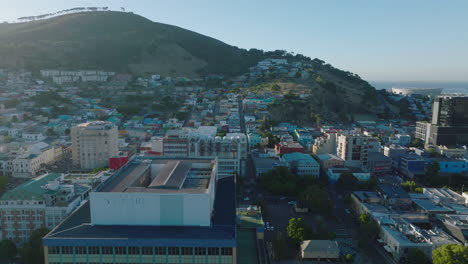 Volar-Sobre-Edificios-Altos-Y-Dirigirse-Hacia-El-Distrito-Residencial-De-Bo-Kaap.-Hileras-De-Casas-Bajas-De-Colores-Y-árboles-En-La-Cima-De-Signal-Hill-En-El-Fondo.-Ciudad-Del-Cabo,-Sudáfrica