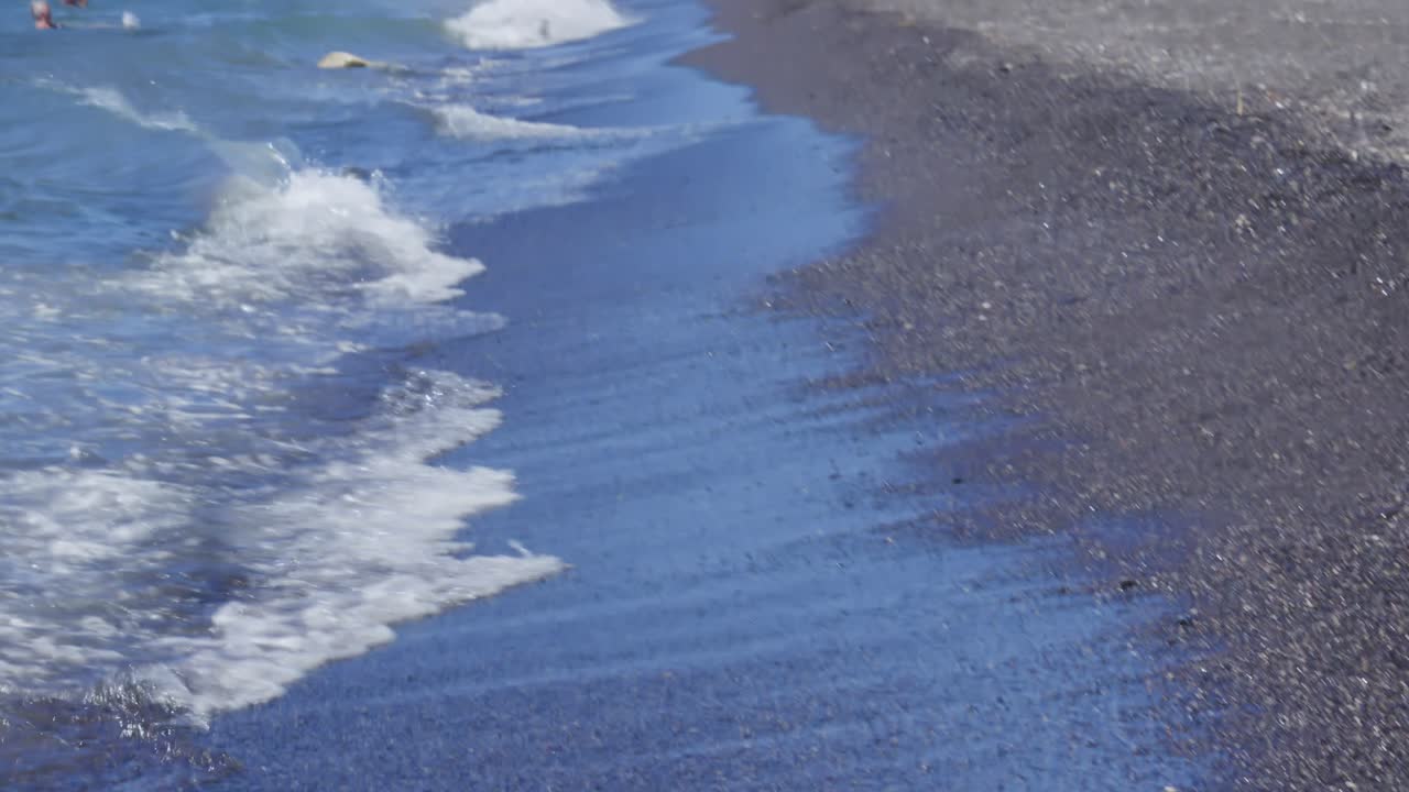 People standing in dark ocean waves crashing along volcano black sand beach coastline in Santorini Greece