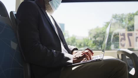 african american senior man wearing face mask using laptop while sitting in the bus