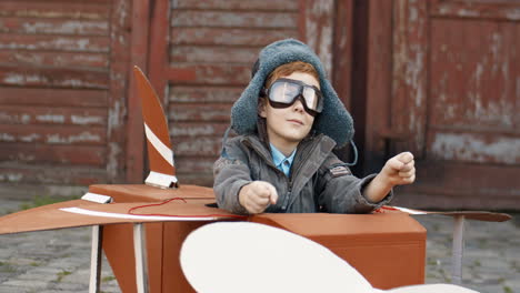 Glücklicher-Kleiner-Junge-Mit-Roten-Haaren-In-Hut-Und-Brille,-Der-Draußen-In-Einem-Hölzernen-Spielzeugmodell-Eines-Flugzeugs-Sitzt-Und-Davon-Träumt,-Flieger-Zu-Sein-1