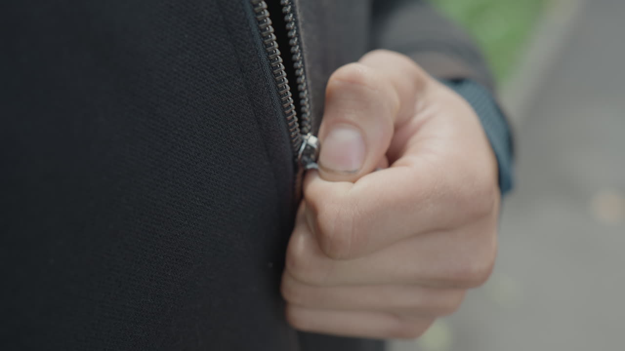 close up tangan mengancingkan ritsleting pada hoodie, pria Kaukasia di jalan setapak taman memegang jaket menahan terpaan angin malam yang sejuk, genggaman protektif dan jari-jari yang mantap, anyaman kain dan slider logam yang terlihat, nyaman dan kuat