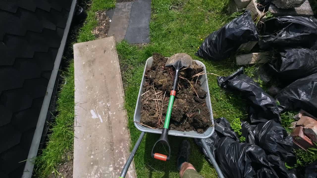 POV of man wheeling a wheelbarrow full of soil and a spade down a garden on a sunny day