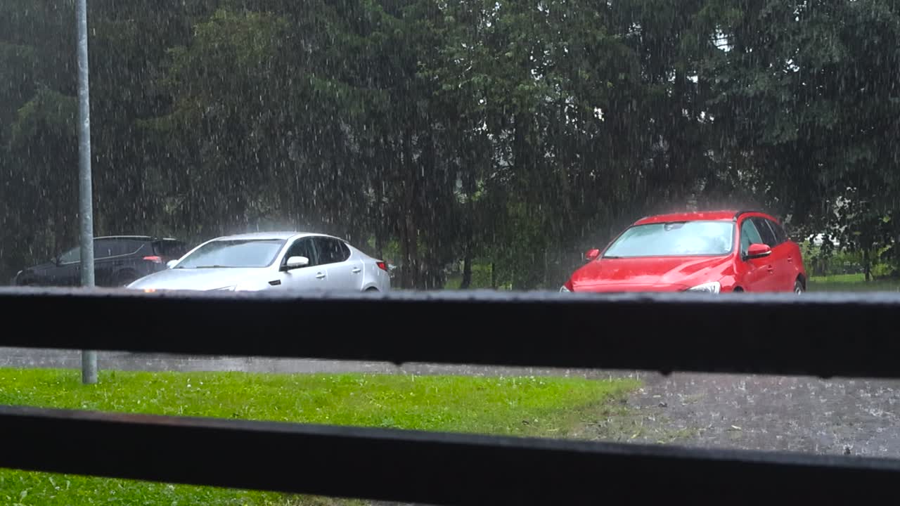 Slow motion video of parked gary and red cars under heavy rainfall during monsoon season at summer or autumn cloudy day with green grass and trees visible in a residential neighbourhood. Wooden rails