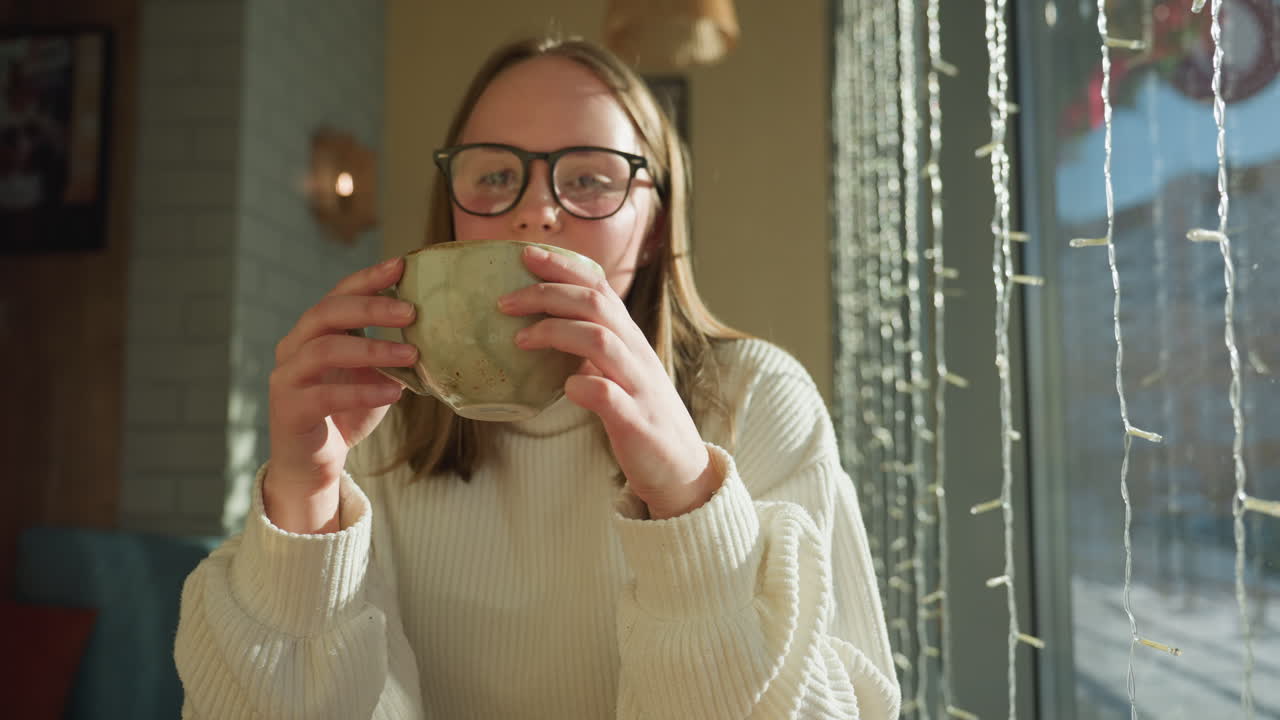 Woman seated beside window holding coffee mug with peaceful expression as snow-covered urban street with parked and passing cars is reflected on glass in soft morning sunlight