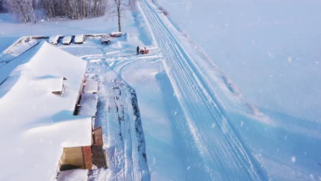 Lonely-person-blowing-snow-near-rural-farm-building-during-snowfall-in-cold-winter